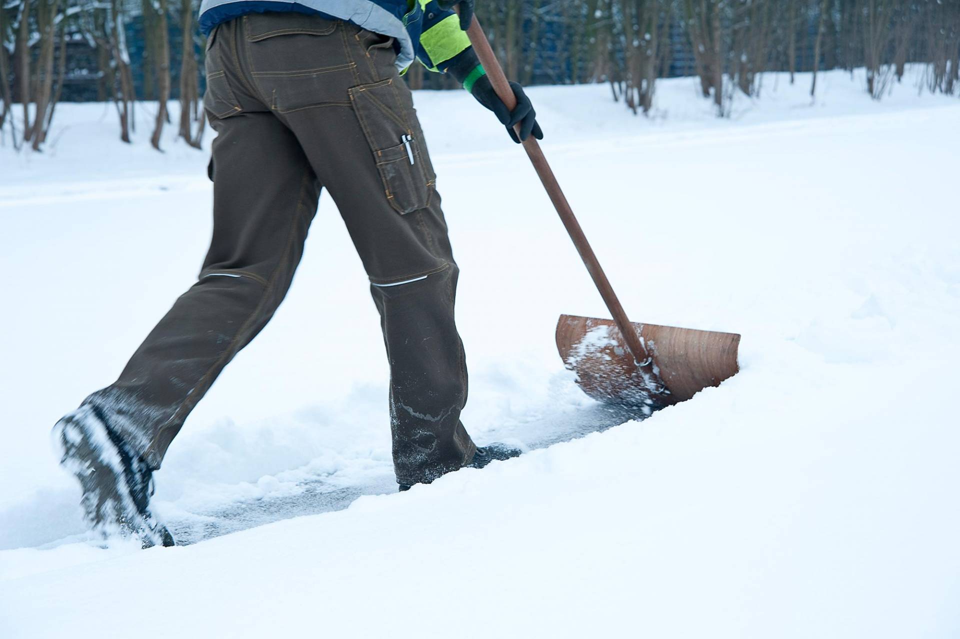 Nahaufnahme eines Winterdienstmitarbeiters in dunkler Arbeitskleidung und Gummistiefeln beim Schneeschippen, zu sehen sind die Beine und eine Schneeschaufel im Einsatz auf verschneitem Untergrund