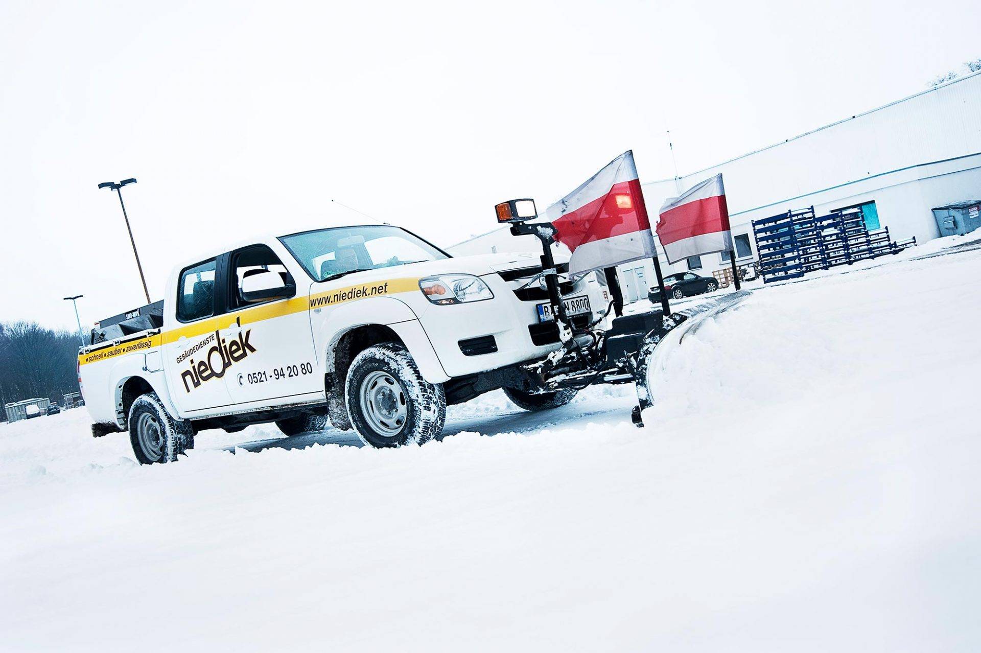 Firmenwagen von niediek.net mit Schneepflug im Wintereinsatz, weißer Transporter mit Firmenwebsite auf der Seite und montiertem Räumschild bei verschneiten Straßenverhältnissen